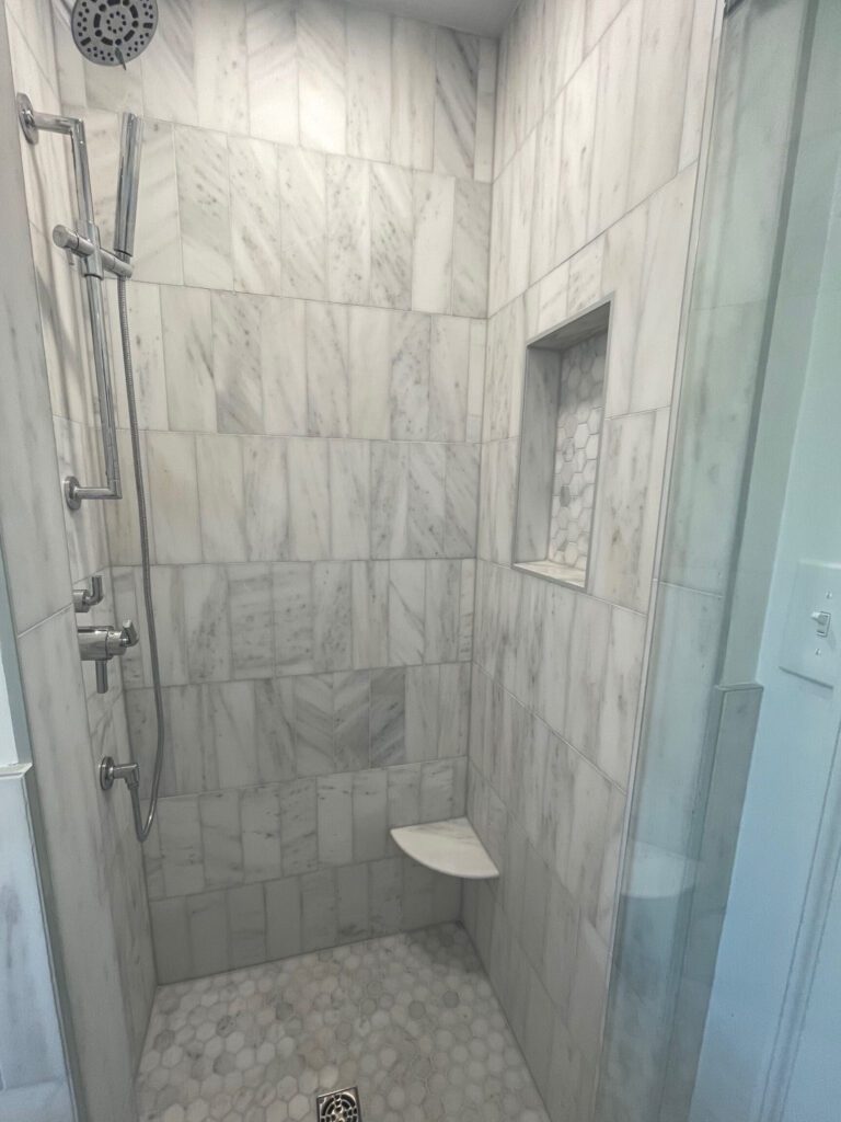The flooring and shower feature polished Bianco Carrara honeycomb tile, while the wainscoting and shower walls are finished in vertical offset Blanco Carrara tile, giving a soft architectural feel.
